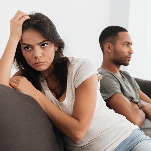 A couple seated on a couch, the female has her back to the male, they both look upset