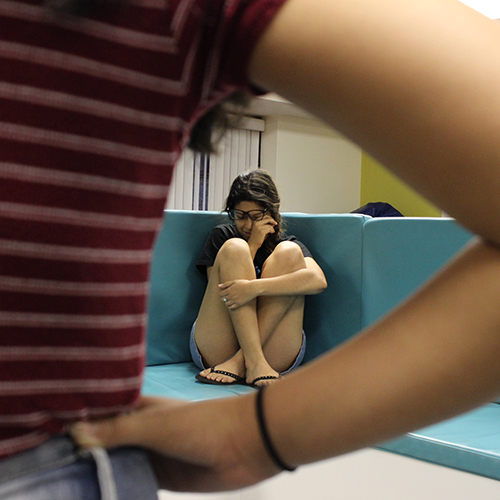 A young woman is cowering in a corner and another young woman is standing with arms akimbo